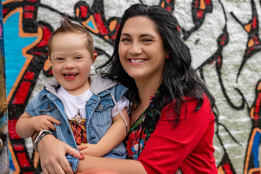 A mum smiles as she holds her young son  in front of a graffiti wall.