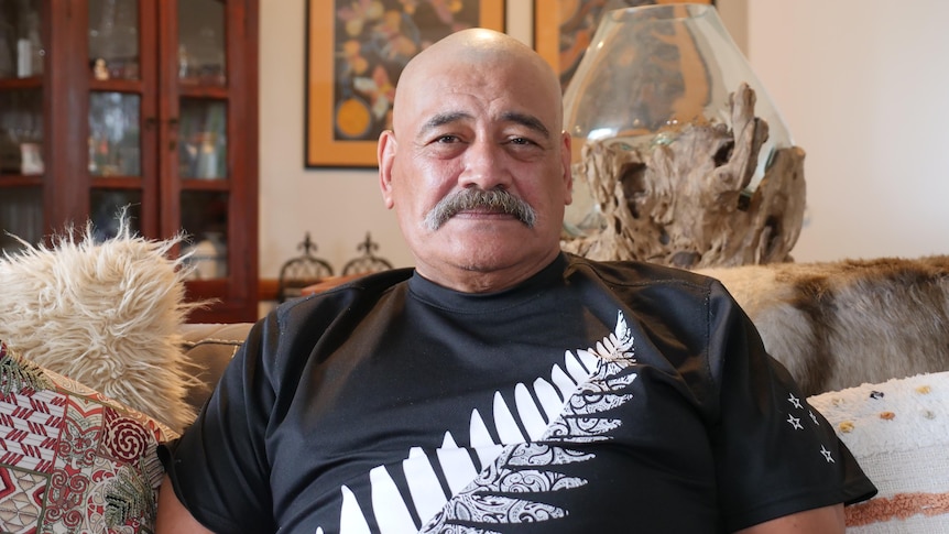 A bald man with a moustache smiles lightly while sitting on a leather couch inside a living room