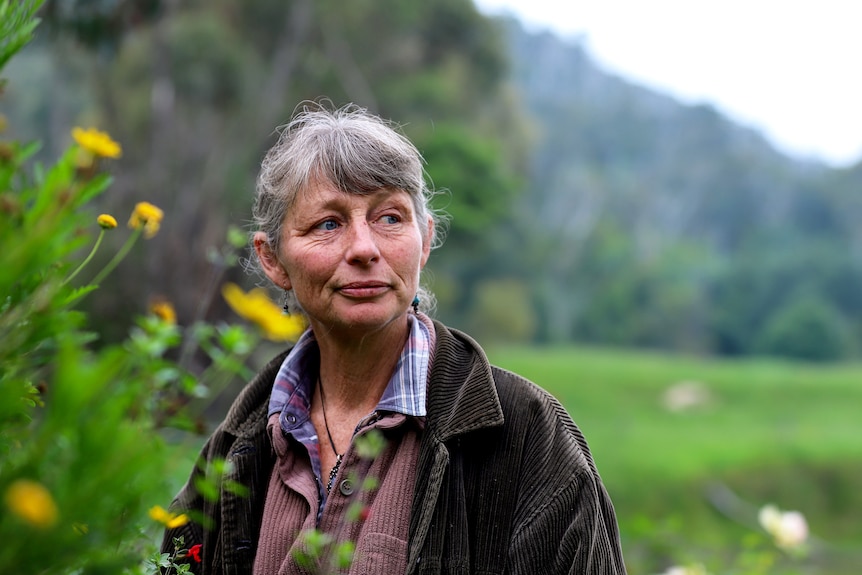 Woman with grey hair wearing brown jacket and shirt stands amid flowers with greenery in background