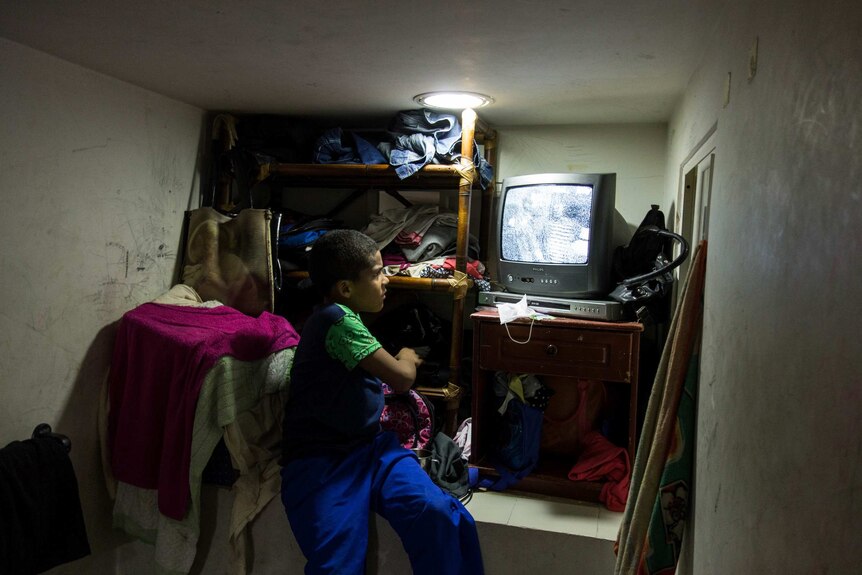 A boy watches tv on an old set in a tiny, messy room.