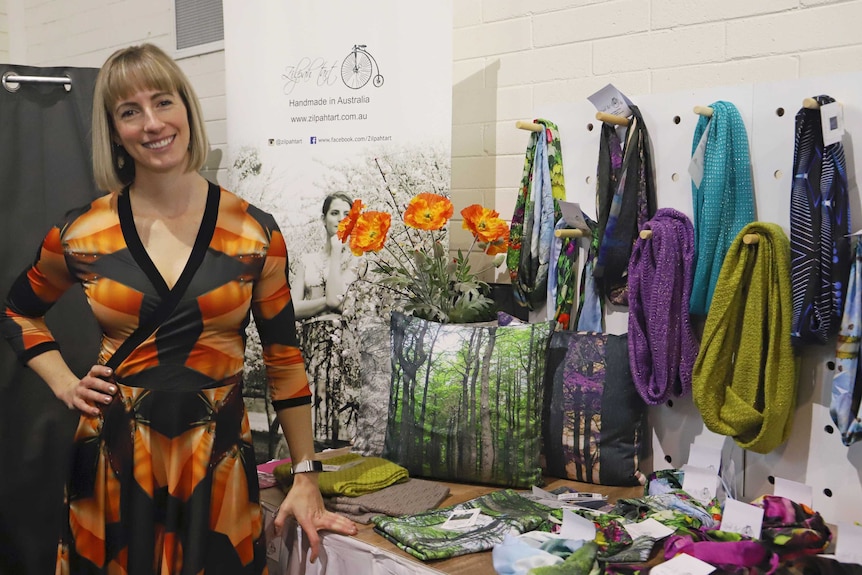 A woman stands in front of a clothing stall with colourful scarves and dresses.