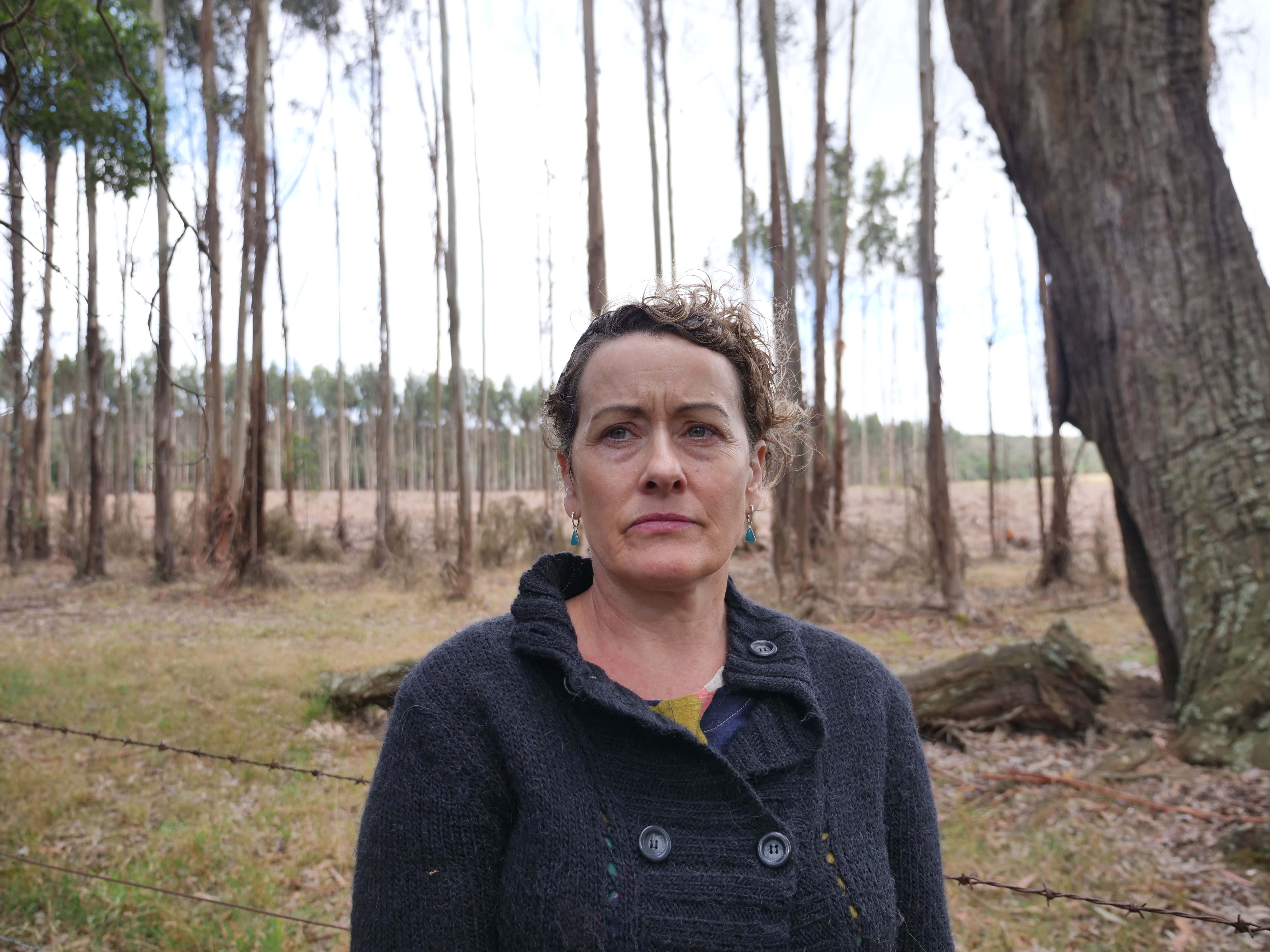 A woman stands in front of a thin row of trees with a cleared paddock behind her.