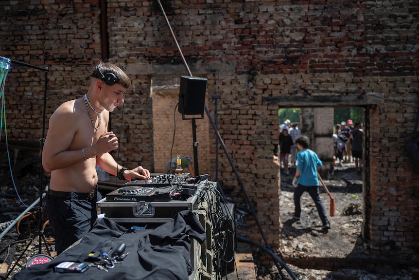 A shirtless man DJs while people in a ruined building clean up bricks and debris 