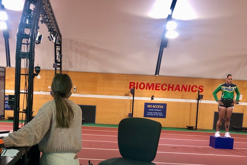 A person sits at a desk looking at the results while an athlete stands on a running track with monitoring cables attached.