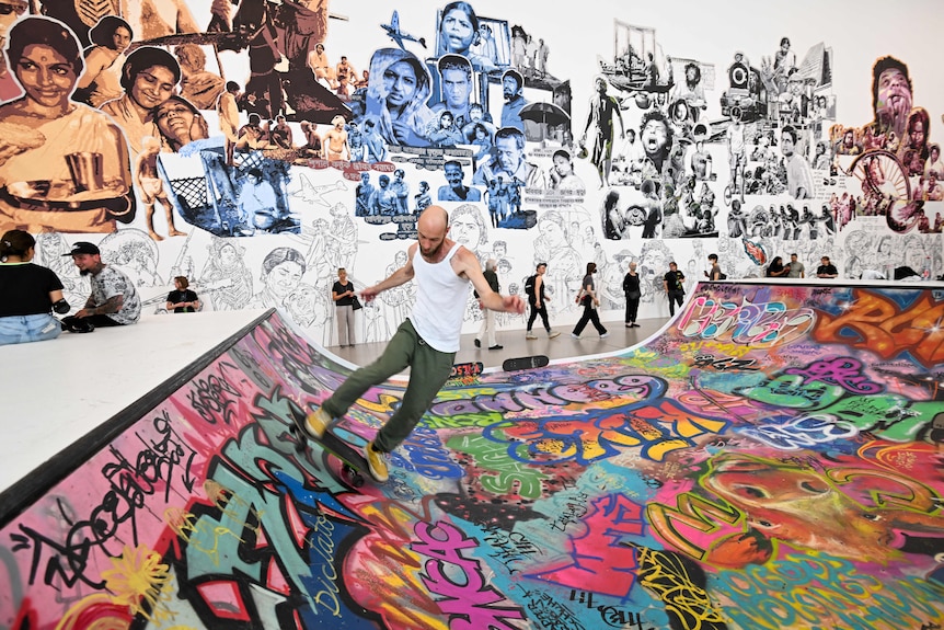 A skater performs on a skateboarding ramp covered in bright graffiti, inside a gallery space.