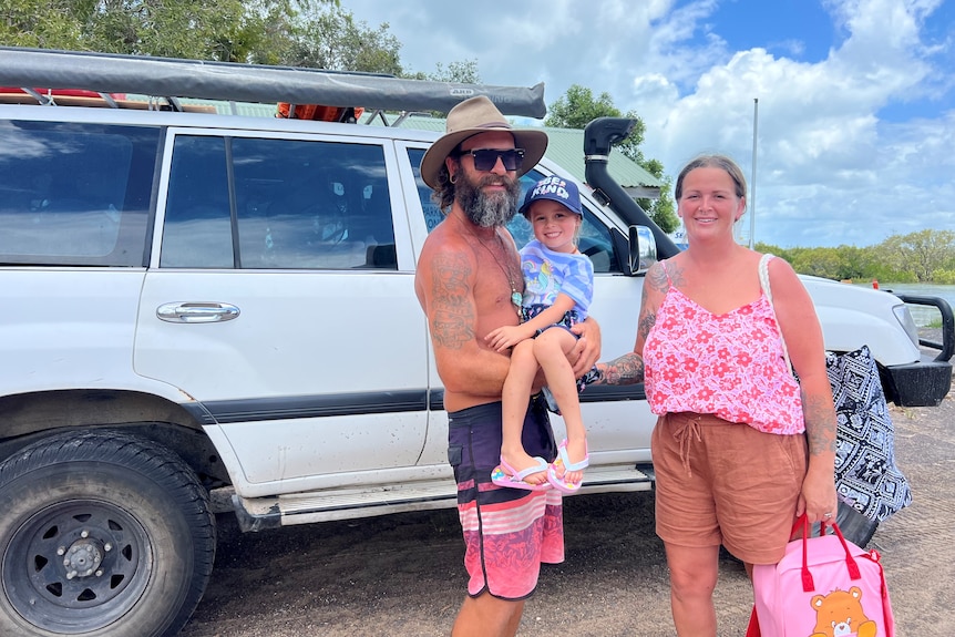 A man carrying a little girl and a woman dressed for the beach stand in front of a 4WD smiling at the camera.