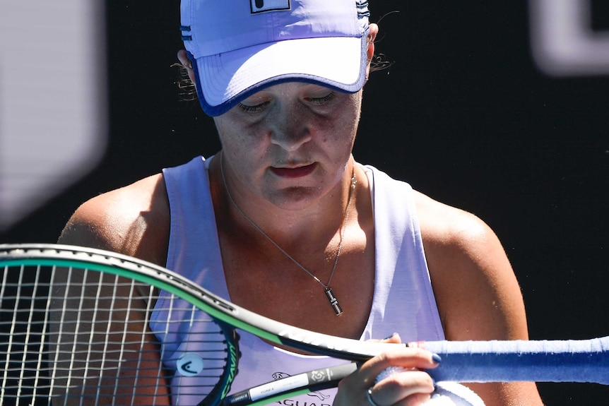 Ash Barty looks down at her racquet which she holds at the top of the handle, horizontal with the floor