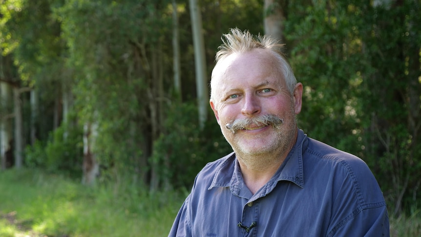 Man with a handlebar moustache and blue shirt standing in front of trees