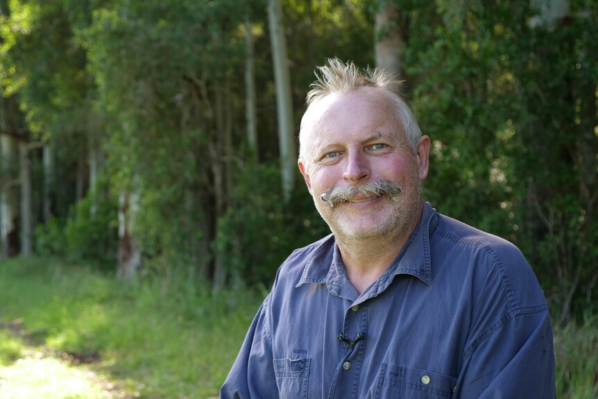 Man with a handlebar moustache and blue shirt standing in front of trees