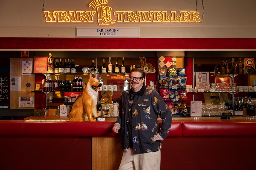 A mustachioed brunette white man wearing a black shirt with animals on it and dark glasses leans against a red leather pub bar.