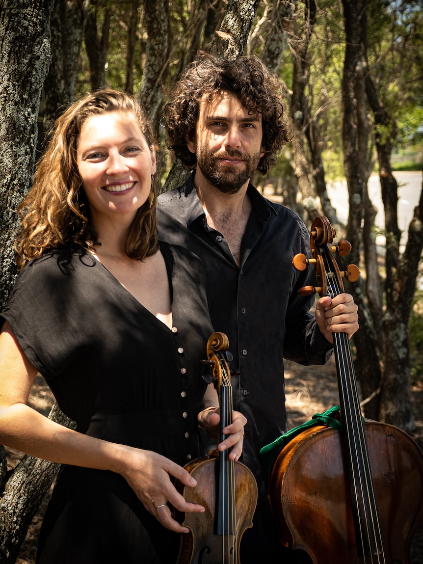 Two people holding instruments and wearing black are standing in a forest They both look happy.