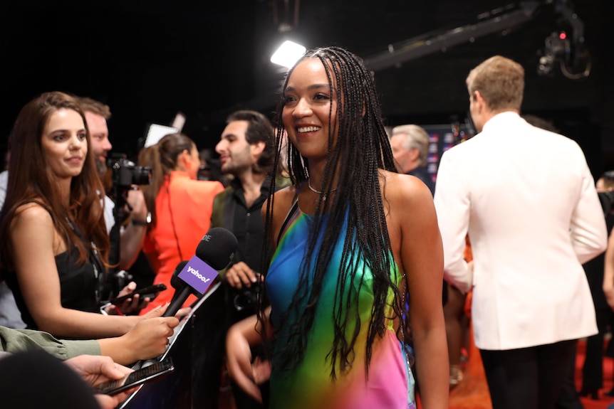 A Black woman in her late 20s with braids and rainbow dress standing on a red carpet, microphone in front of her