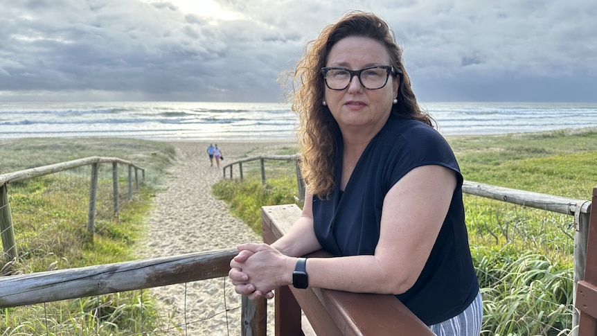 A slightly smiling middle-aged woman leans against a wooden post, in front of a beach. Dark curly hair, glasses, blue top.