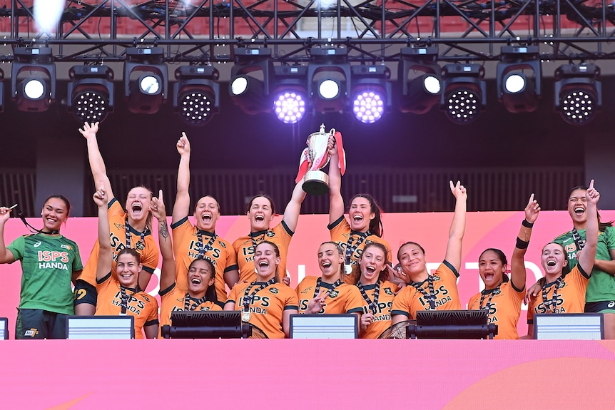 The Australian women's rugby sevens team lifts a trophy and has arms in air cheering.