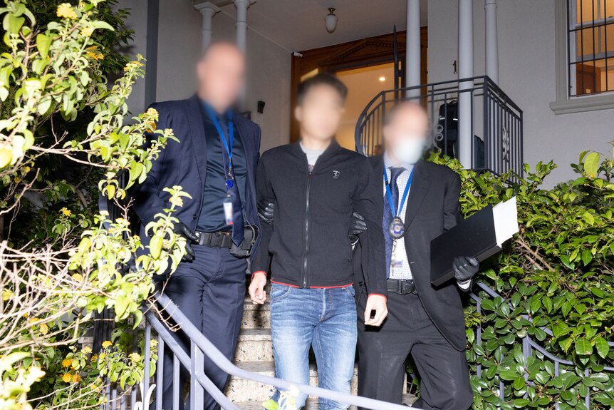 A man with a blurred face flanked by detectives in suits