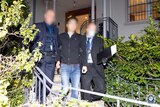 A man with a blurred face flanked by detectives in suits