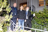 A man with a blurred face flanked by detectives in suits