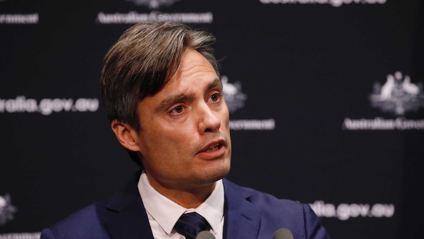 Man with brown hair in front of a black background in the middle of speaking.