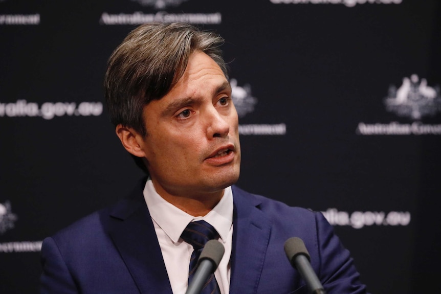 Man with brown hair in front of a black background in the middle of speaking.