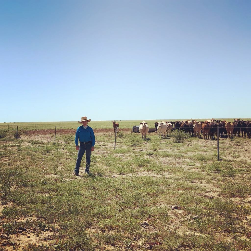 Woman dressed in farmer atire of jeans, button up blue workshirt and cowgirl hat stands in front of paddock of cows with blue sk