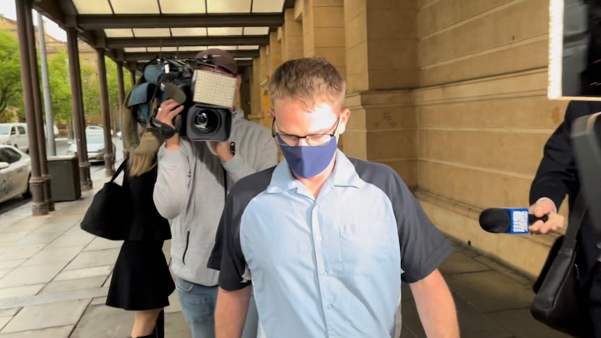 A man wearing a light blue buttoned shirt with navy sleeves and a navy mask walks with cameras behind him