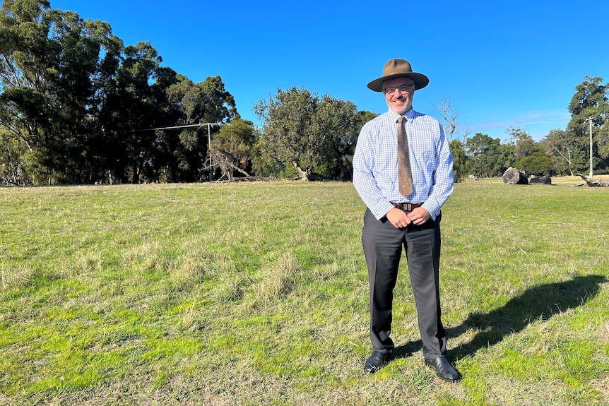 A man in a suit and hat stands in a green grass paddock. 