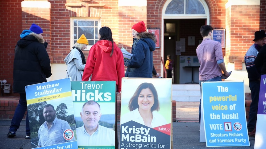 Candidate promotional signs and people standing outside a polling booth.