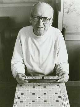 A black and white photo of a man in glasses in front of a Scrabble board, holding tiles spelling Alfred Butts