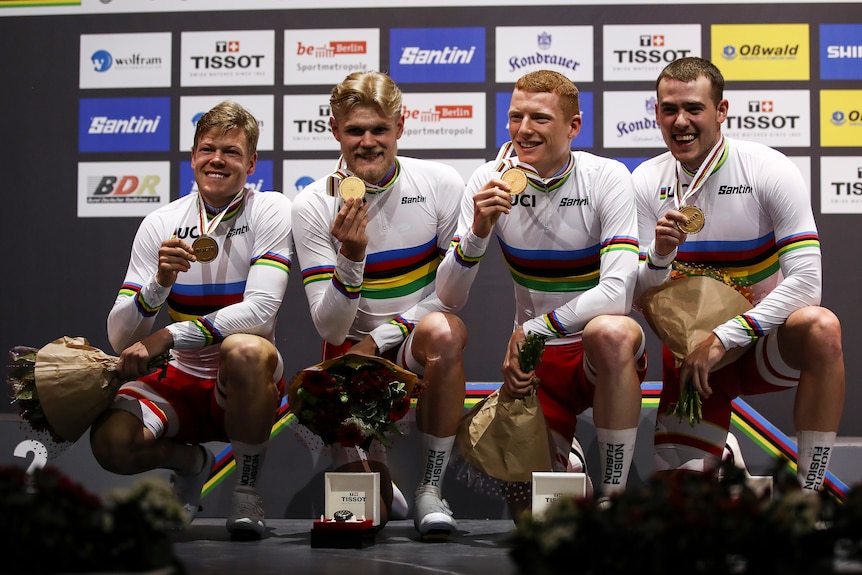 Four men kneel down holding onto gold medals