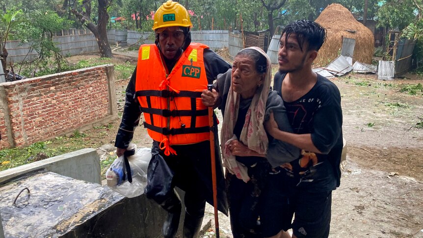 Rescue workers help an elderly woman to reach a makeshift shelter, guiding her up stairs as rain falls in the background.