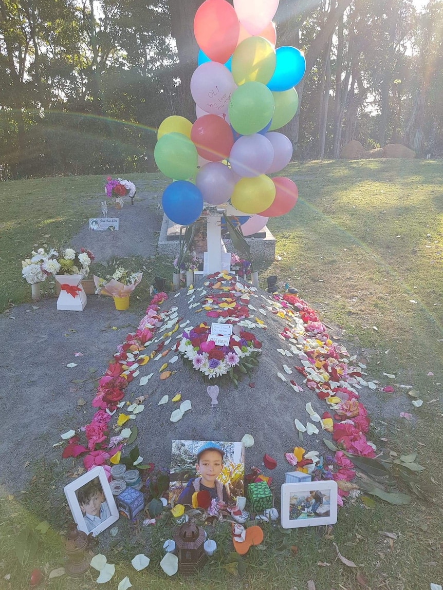 A child's grave with a column of balloons and a ring of flowers, fronted by a photo of a young boy.