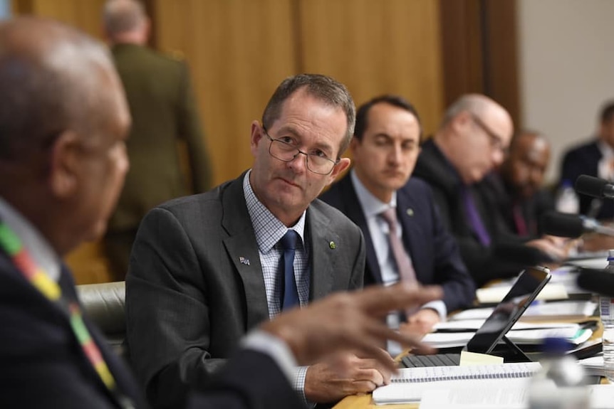 A man in a suit speaks to other similarly attired men at a conference table.