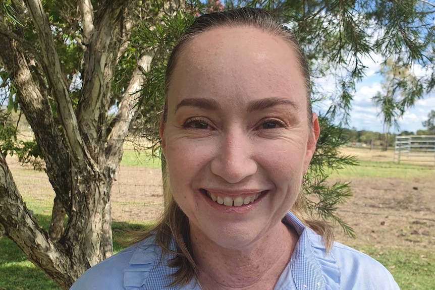 A woman wearing a blue button up shirt with ruffles smiles. She has brown hair in a pony tail.