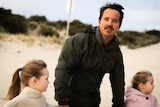 A man with dark hair and a moustache stands on a beach with his two young daughters 