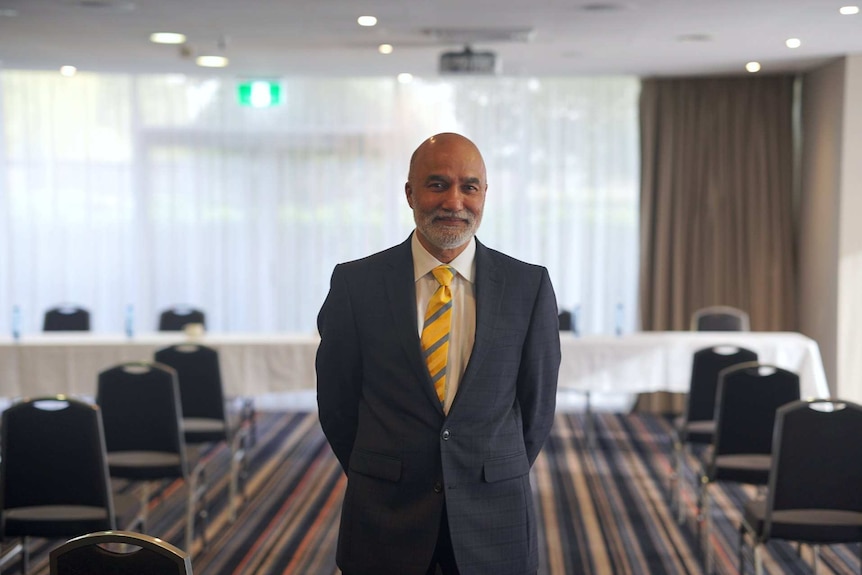 A smiling man in a suit and gold tie stands with his hands behind his back in a function room.