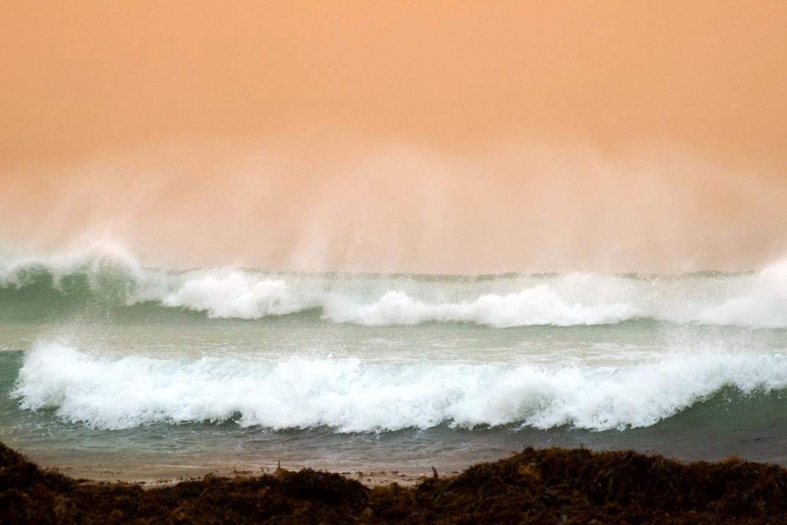 Large waves crash sending spray into the air backlit by an orange dust-filled sky.