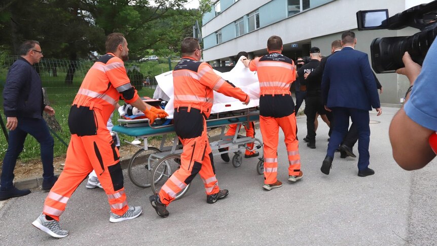 Several people wheel a stretcher in to a large building. The person on the stretcher is obscured by white sheets.