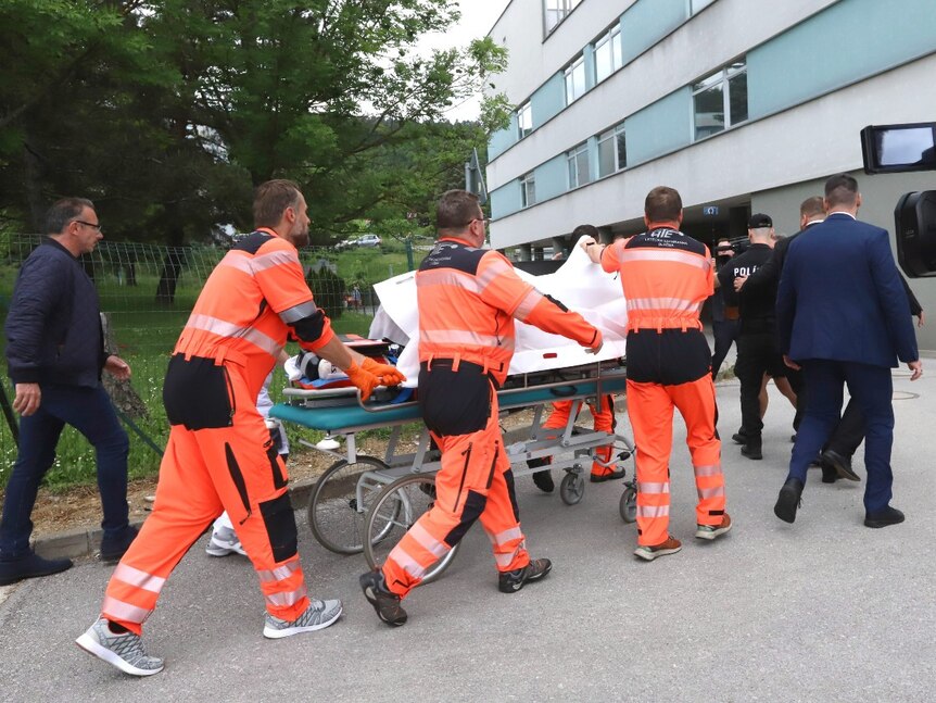 Several people wheel a stretcher in to a large building. The person on the stretcher is obscured by white sheets.