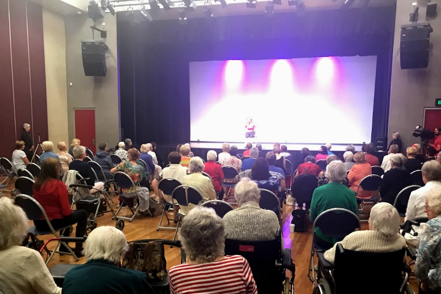 About 100 seniors sitting on chairs in a large theatre, they're looking at the stage where a woman is talking on stage.