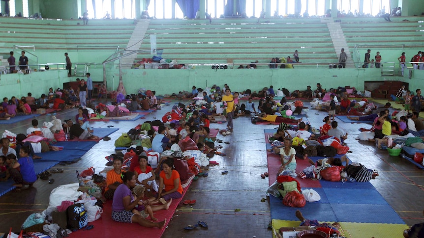 Tens of villagers sit and lay on mats in a sports centre.
