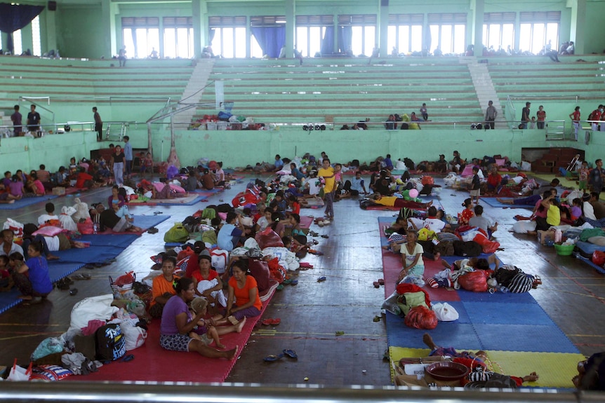 Tens of villagers sit and lay on mats in a sports centre.