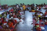 Tens of villagers sit and lay on mats in a sports centre.