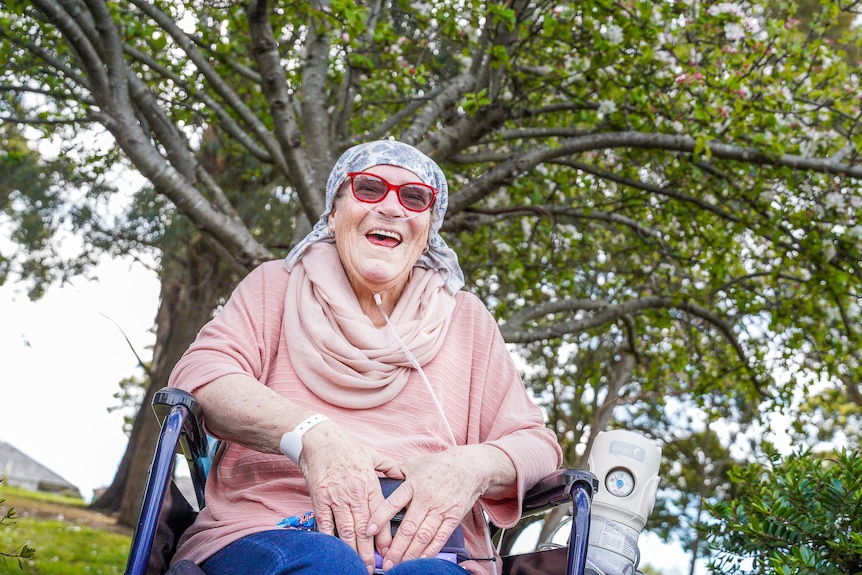 A woman with a shaved head wearing red sunglasses laughs in a wheelchair under a shady tree.