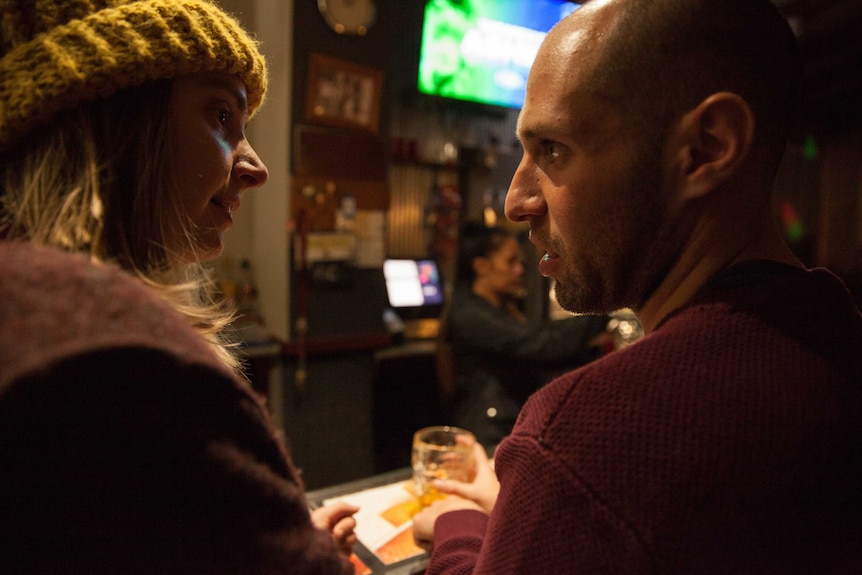 Josh and his girlfriend lock eyes, his hand curled around a drink, as they stand at a bar.