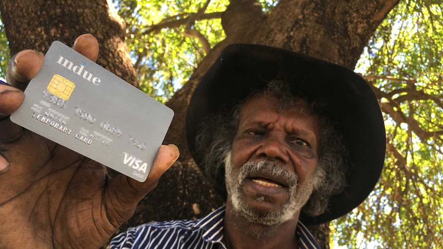 A serious, bearded Indigenous man wears a hat, blue and white striped shirt, stands under a tree, holds a grey visa card. 