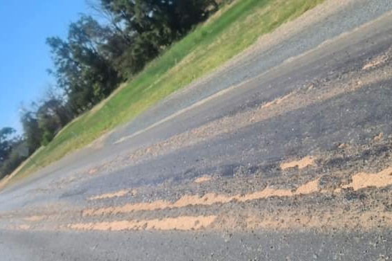 A bitumen road showing bare dirt where surface has eroded, with green strip and trees along edge of road.