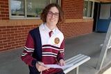 A student in a maroon, white and blue uniform, wearing glasses and with long hair, smiles outside a school building.