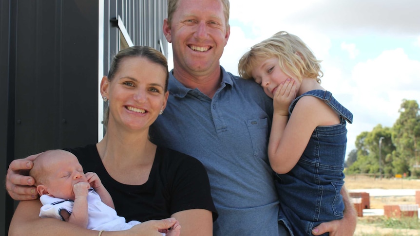 A man and woman stand in front of a shed, with hay bales in background. They are are holding a newborn baby and little girl