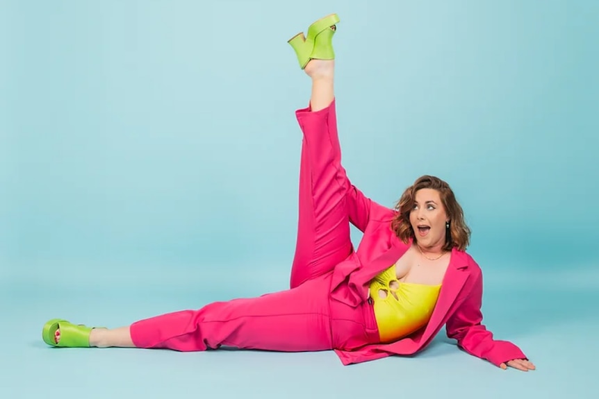 A woman in a bright pink suit and bright green high heels lies on the ground with one leg in the air