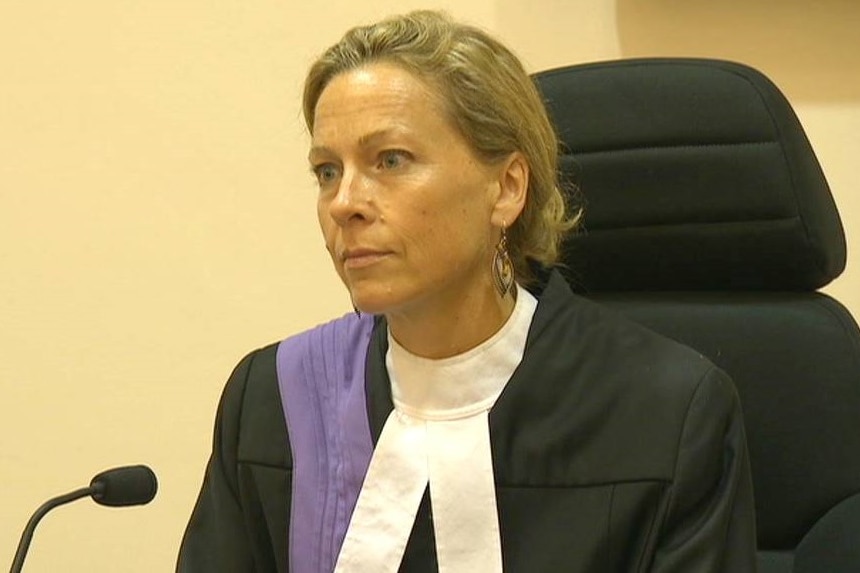 A woman with short blonde hair, wearing a black and white judge's robe, sits at a desk with a microphone